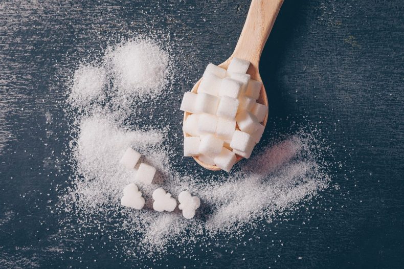 Some white sugar in a spoon on dark background, flat lay.